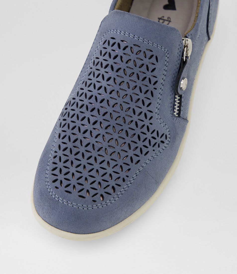 A close-up of a slip-on shoe in dusty blue suede with geometric laser-cut perforations on the toe box, a side zipper with silver metal hardware, and a cream-colored sole.